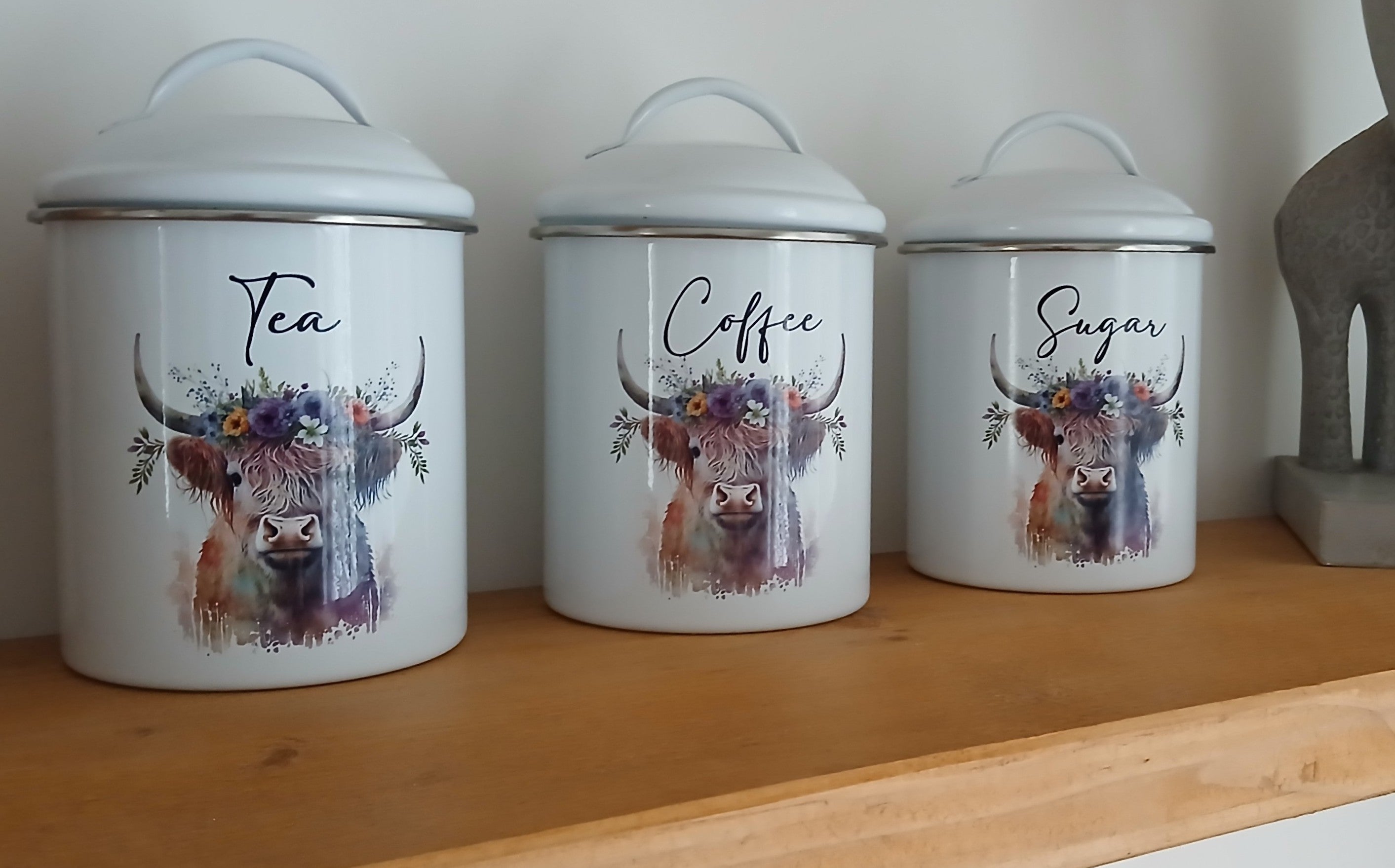 Three white canisters with cow designs labeled 'Tea', 'Coffee', and 'Sugar' on a wooden shelf.